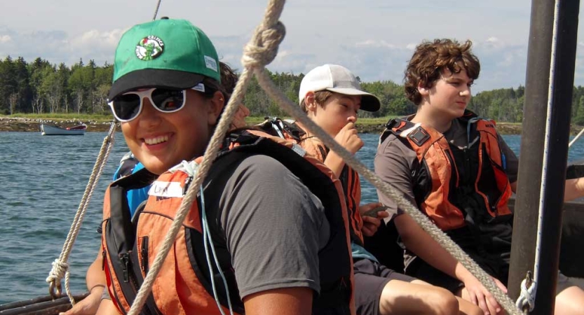 Three teens wearing life jackets sit on a sailboat 